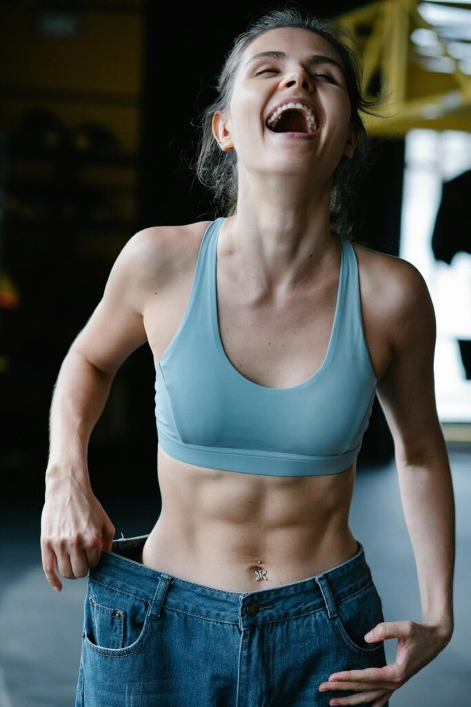 pexels-photo-7991921-7991921 Fit young woman happily shows off weight-loss success by wearing oversized jeans in a gym.